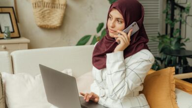 A Muslim woman in a hijab working remotely, balancing a phone call and laptop in a home office setting.