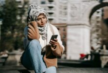 Stylish young Arab female in casual warm outfit and traditional hijab resting on city square and messaging on mobile phone on sunny day