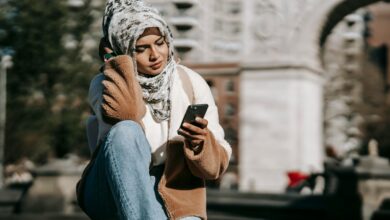 Stylish young Arab female in casual warm outfit and traditional hijab resting on city square and messaging on mobile phone on sunny day