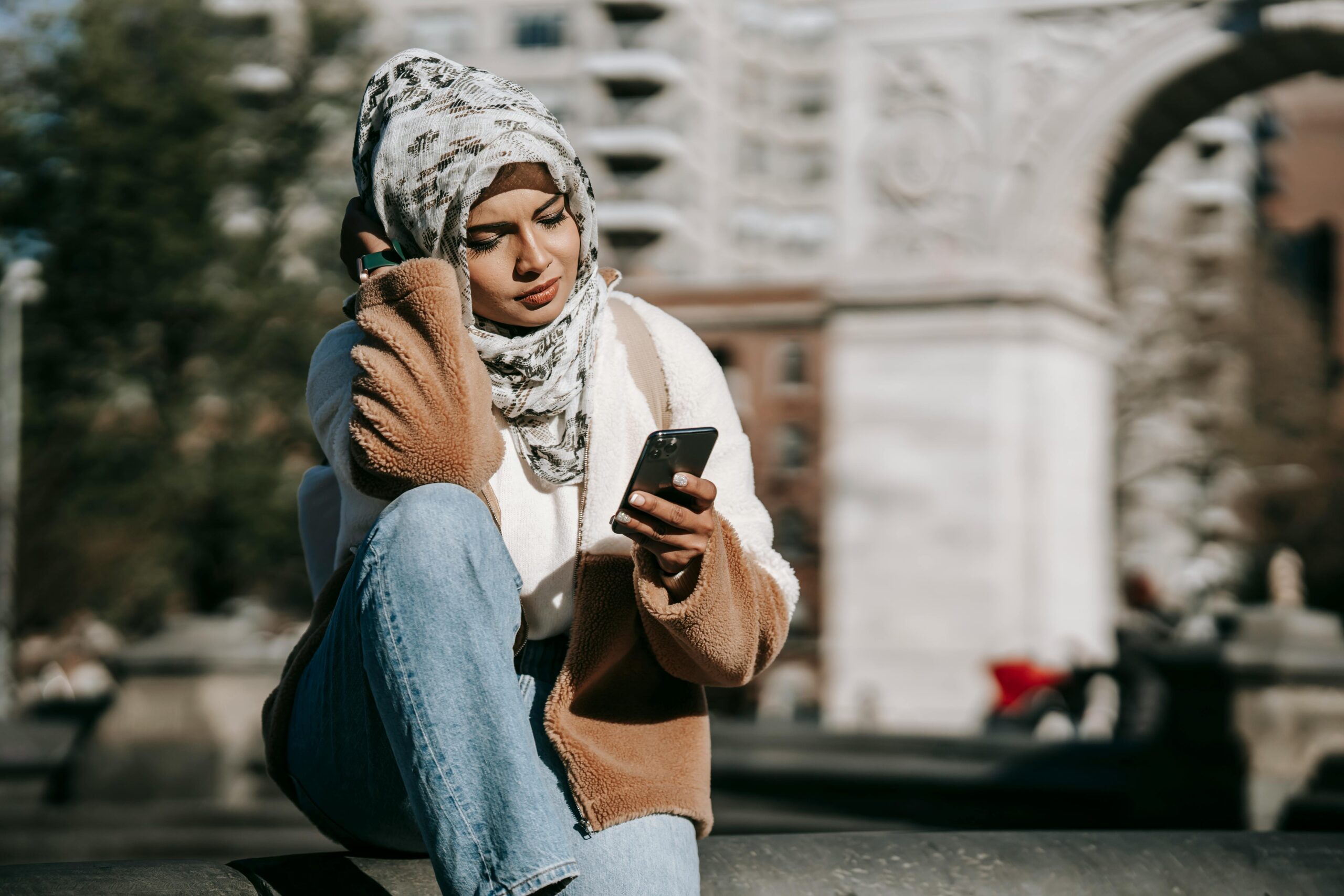 Stylish young Arab female in casual warm outfit and traditional hijab resting on city square and messaging on mobile phone on sunny day
