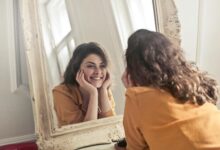 A cheerful woman smiles at her reflection in a vintage-style mirror, exuding positivity and warmth.